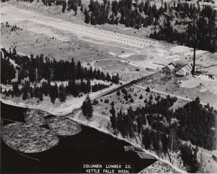 Black and white aerial photograph of buildings on the shore of a lake. 