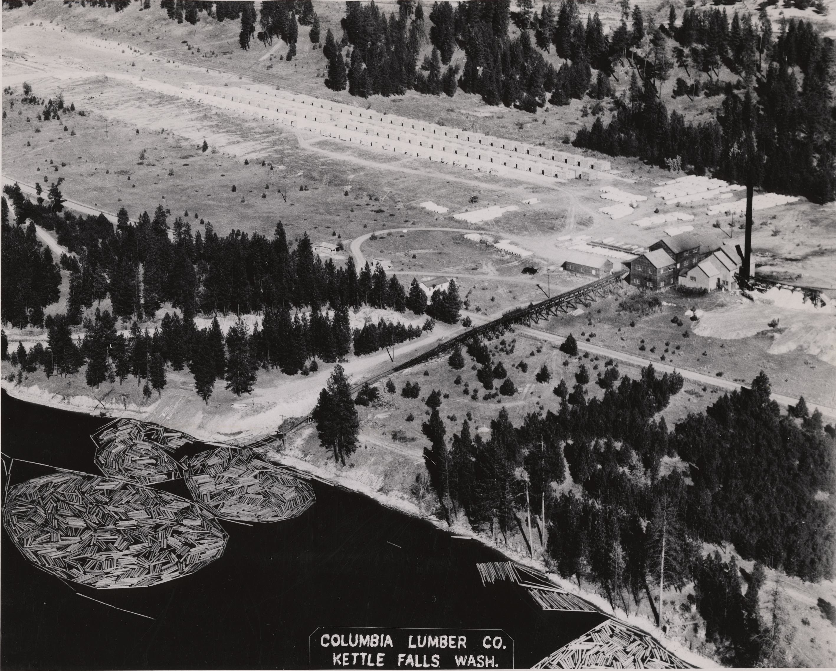 Black and white aerial photograph of buildings on the shore of a lake. 