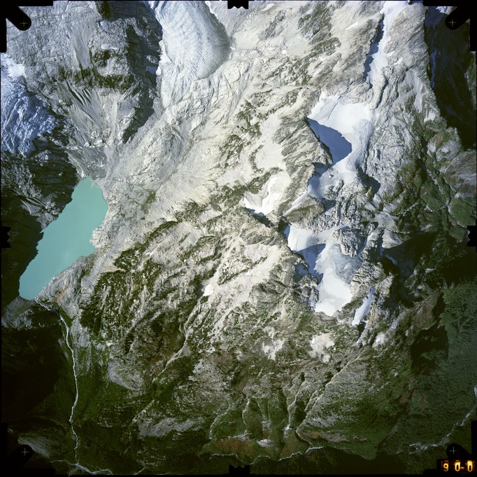 Color aerial photograph of glacier, snowfields, and alpine lake on mountain peak with forested valleys below.