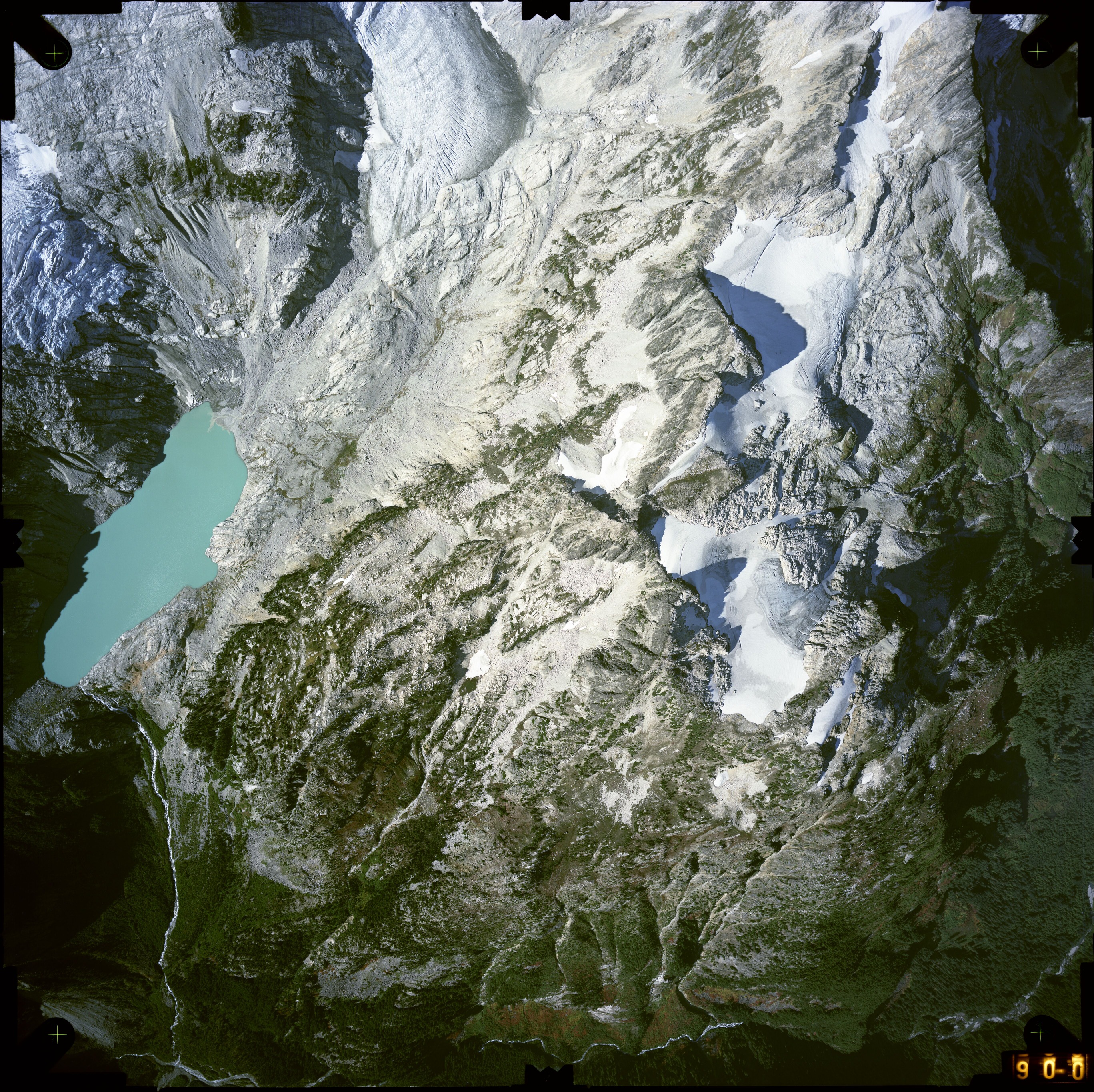 Color aerial photograph of glacier, snowfields, and alpine lake on mountain peak with forested valleys below.