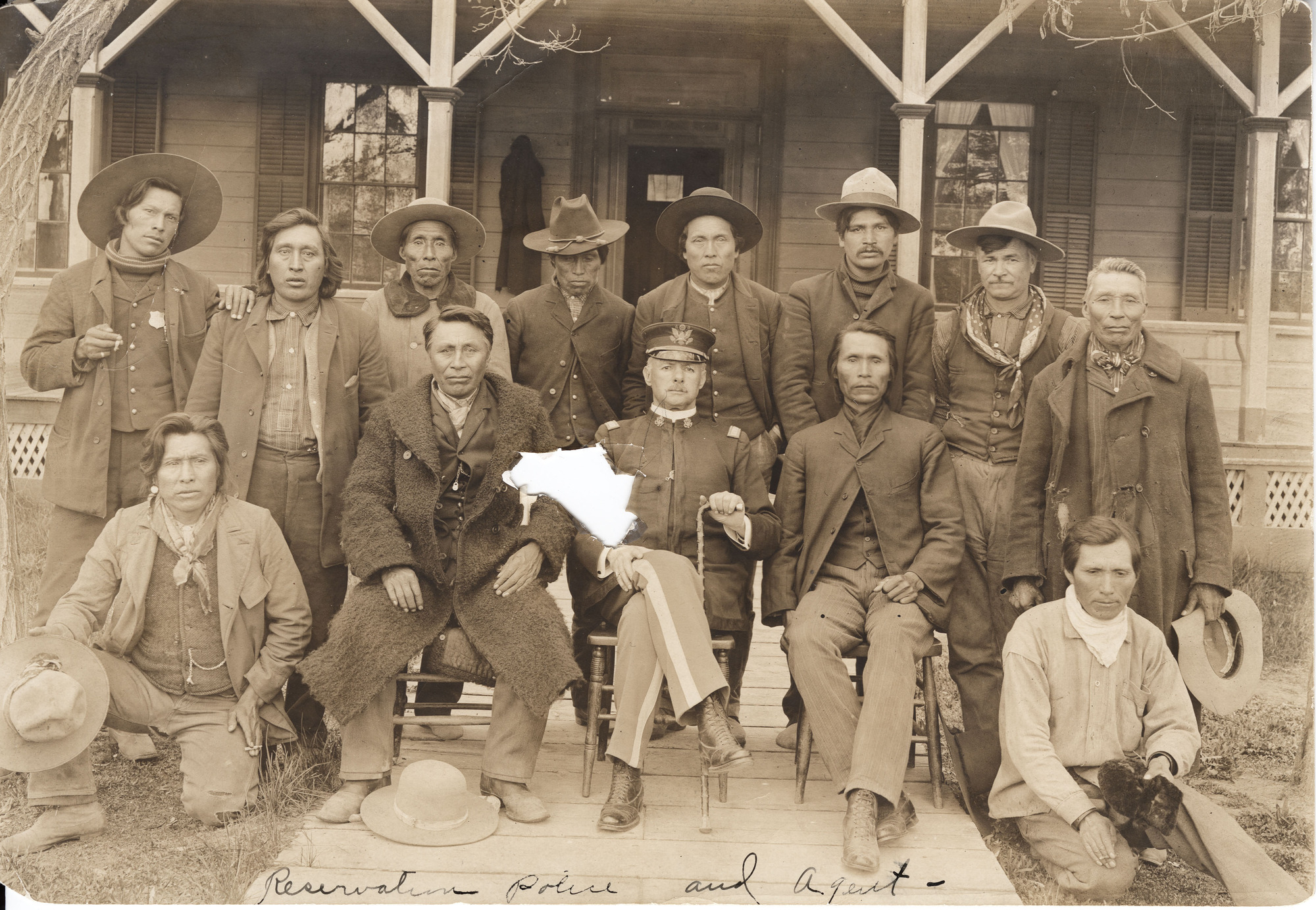Black and white photograph of American Indians in western clothes sitting and standing for a photograph with a soldier in the middle