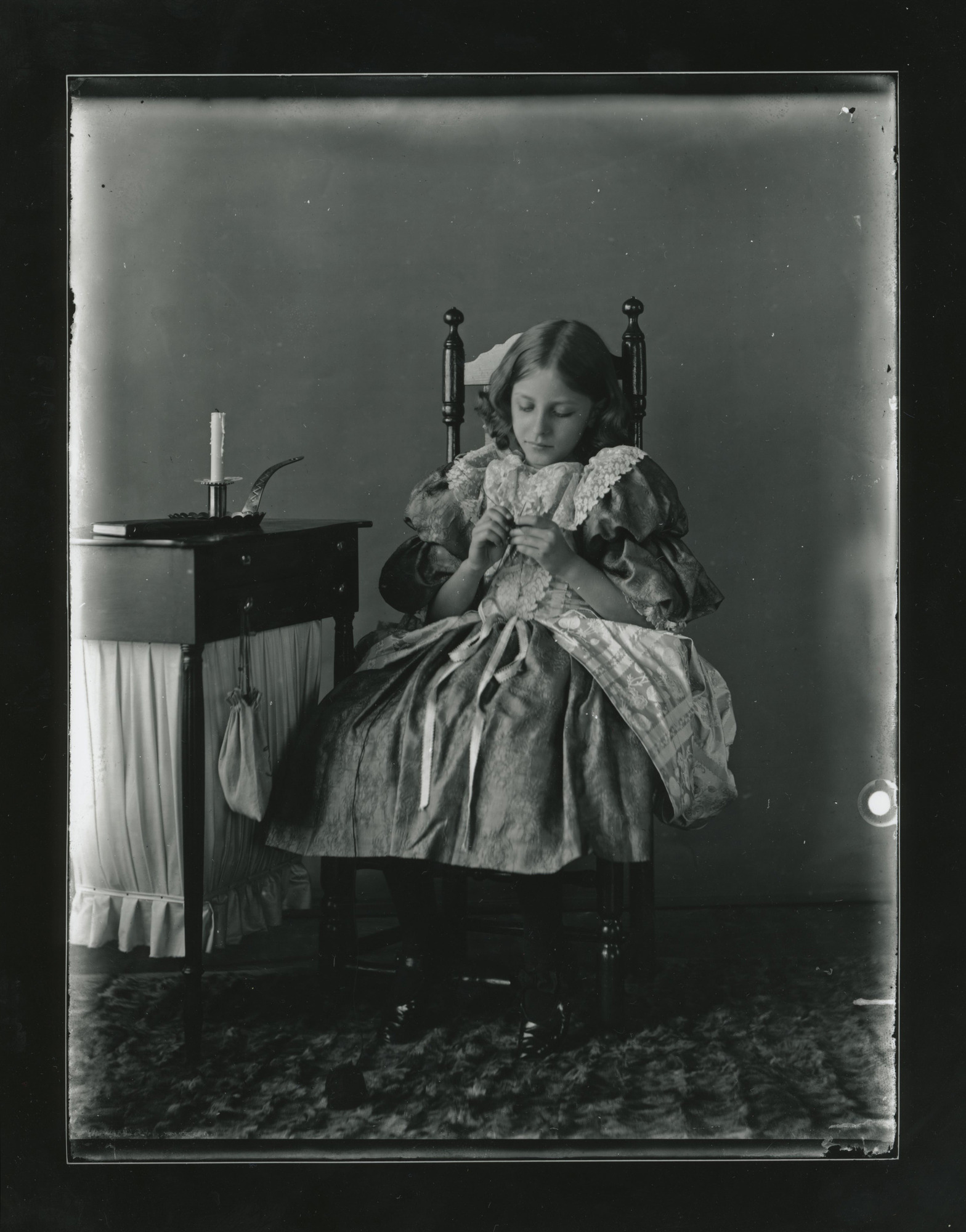 A young white girl sits in a chair and knits. Ball of yarn on floor. Side table with a book and a candle on left.