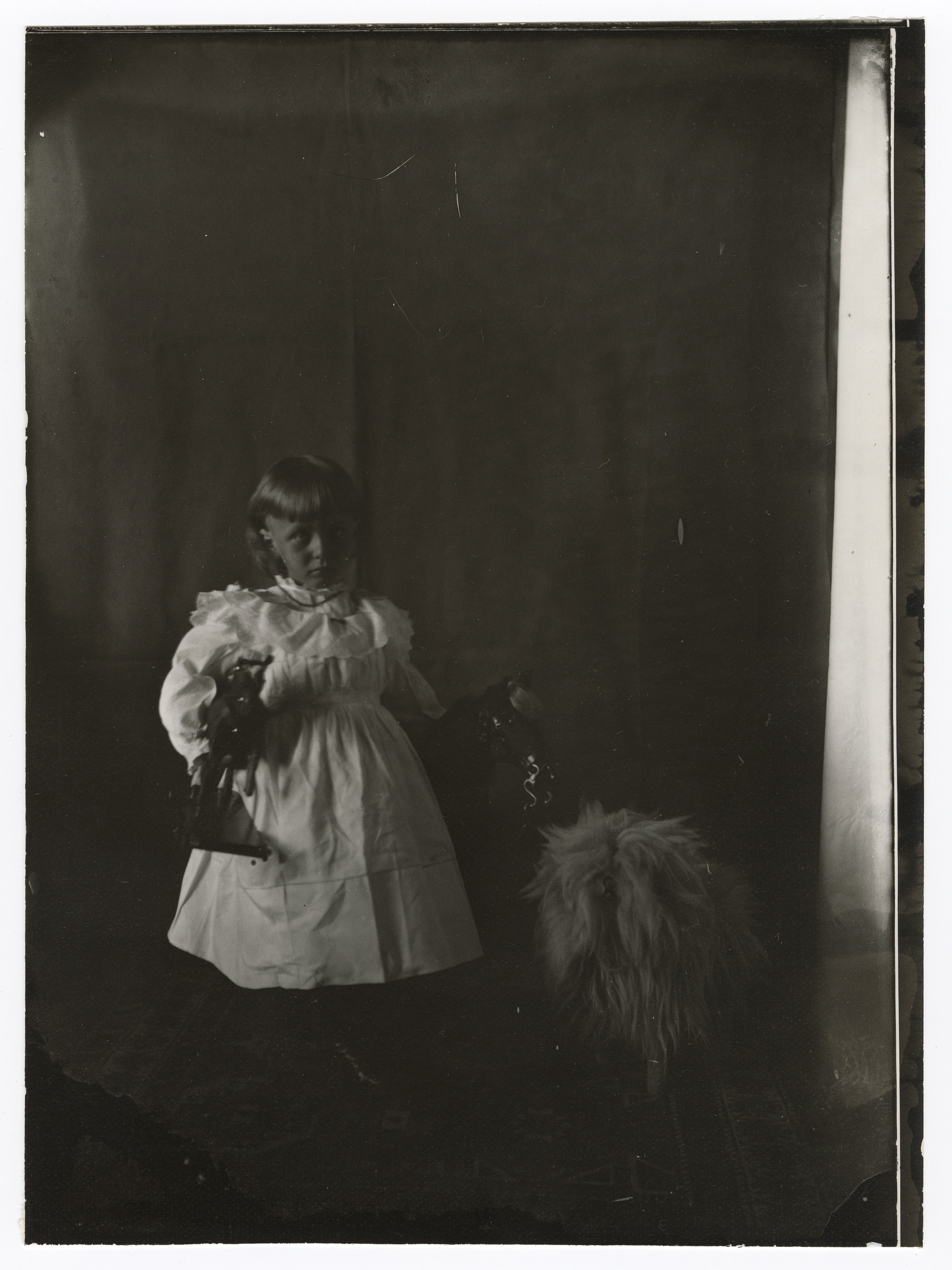 A young white girl in a white dress stands before a rocking horse and holds a statue of a horse in her right hand. Small, shaggy toy dog next to her.