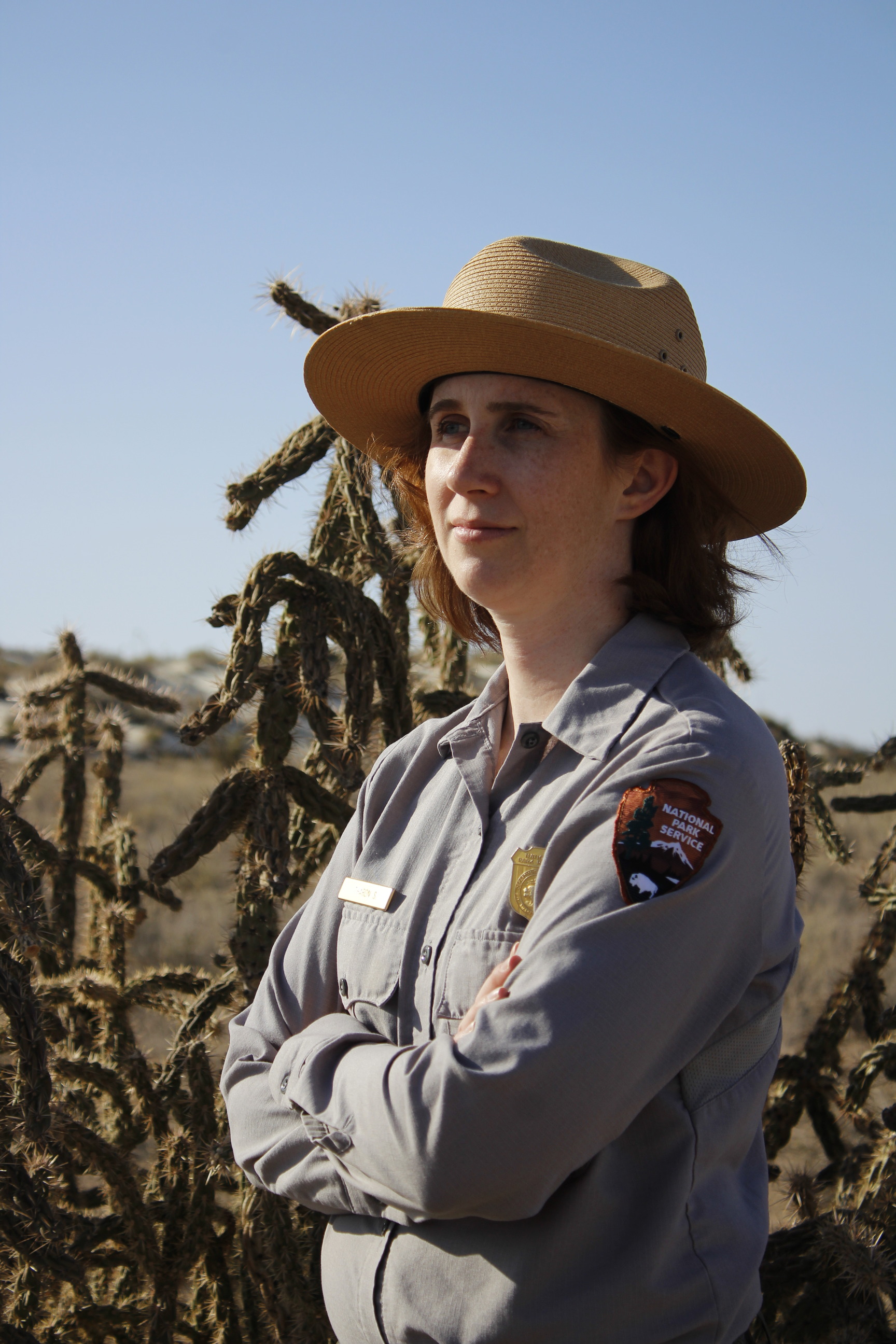 A woman wearing the national park service uniform poses for a photo on the border of the White Sands dune field. Cacti and desert shrubbery extend into the distance meeting the base of white sands dunes. The dunes are freckled with shrubbery and meet a clear light blue sky on the horizon.