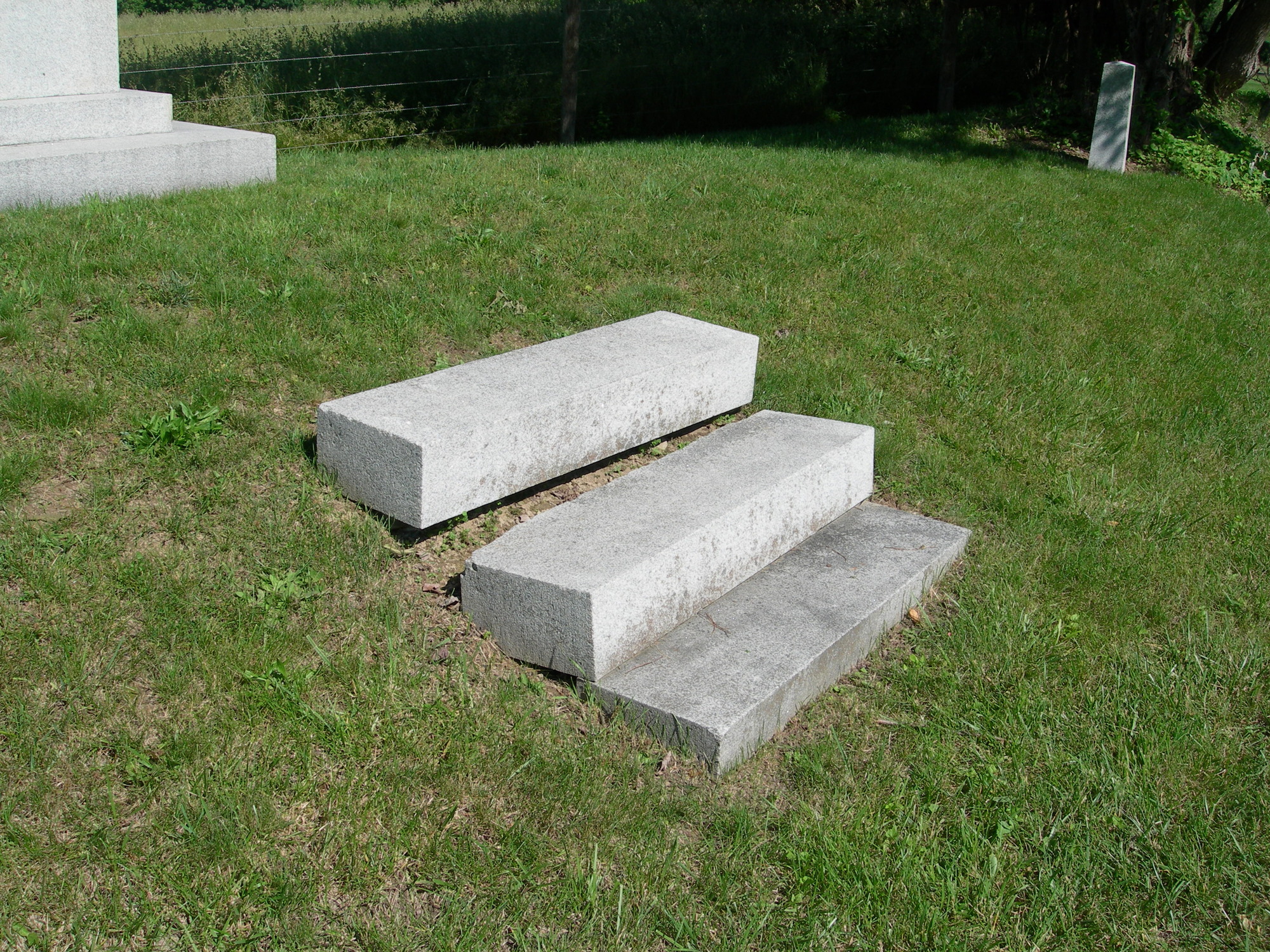 Three granite steps sit in a small hillside. The top step is separated by several inches from the lower two steps and grass is visible between them.