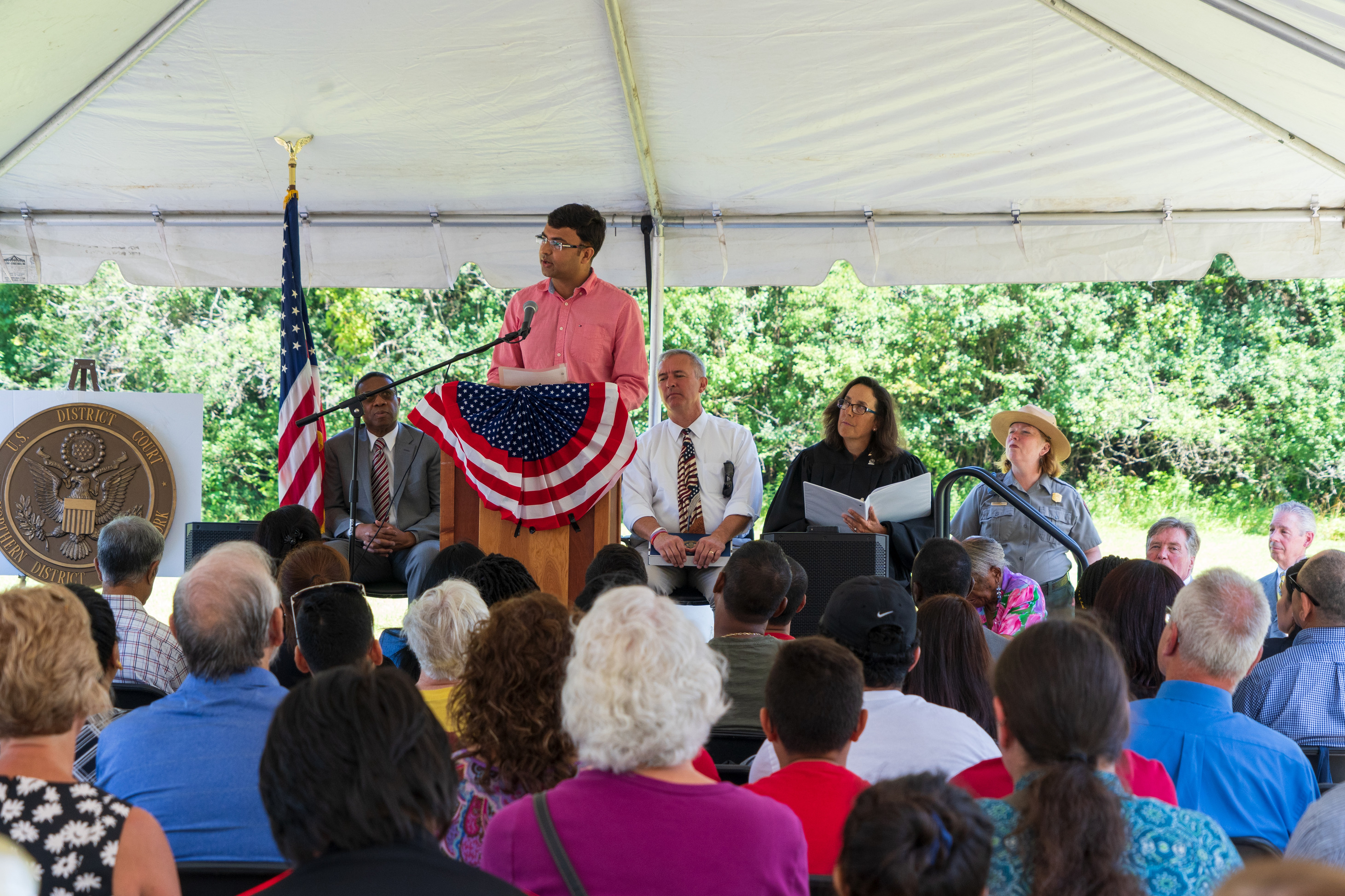 People sit under a tent and listen to a woman speak at a podium.