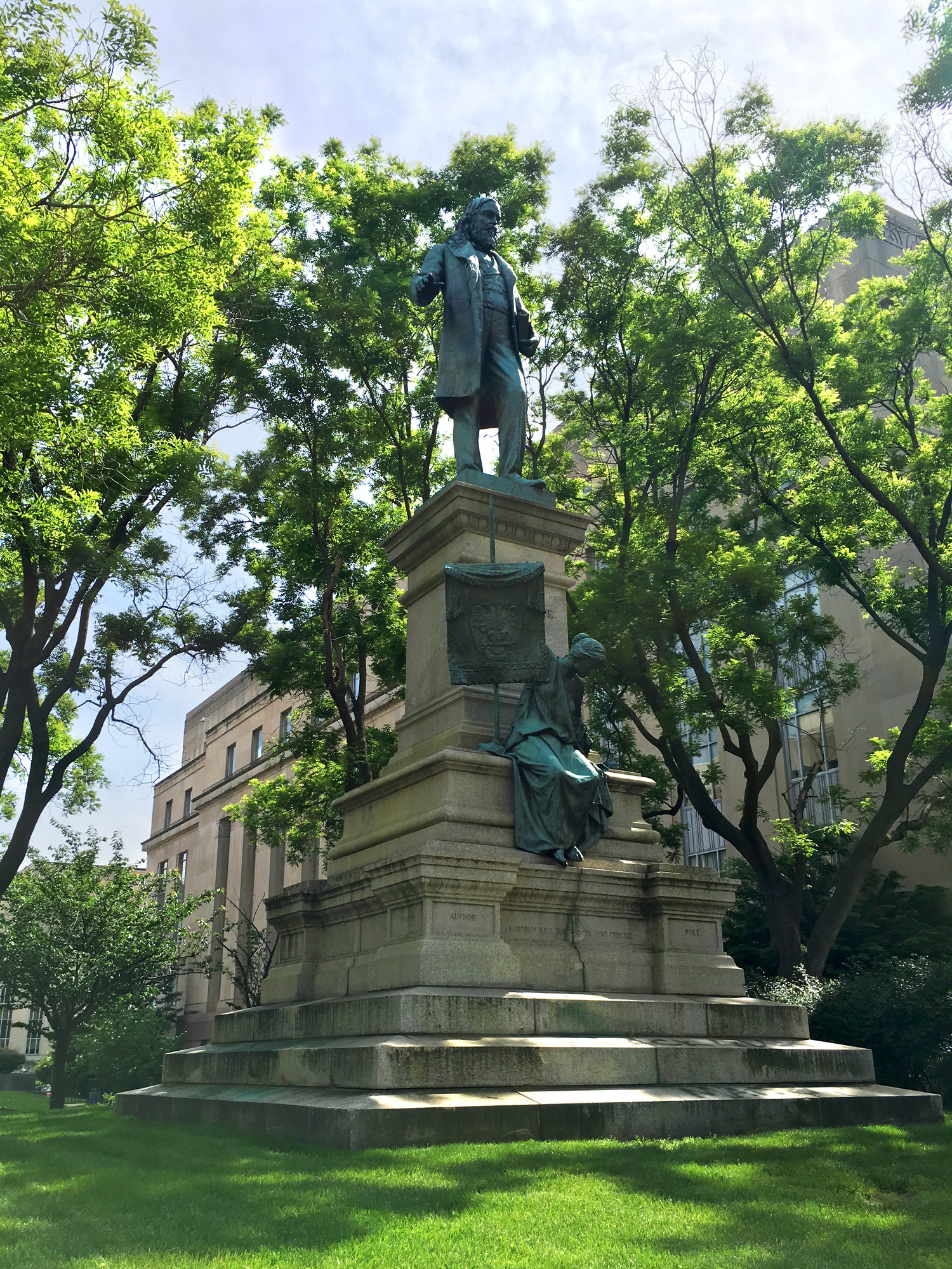 A multi-tiered stone base with a greenish bronze figure of a man in a long coat standing at the top. A female bronze figure sits about halfway of the base. 