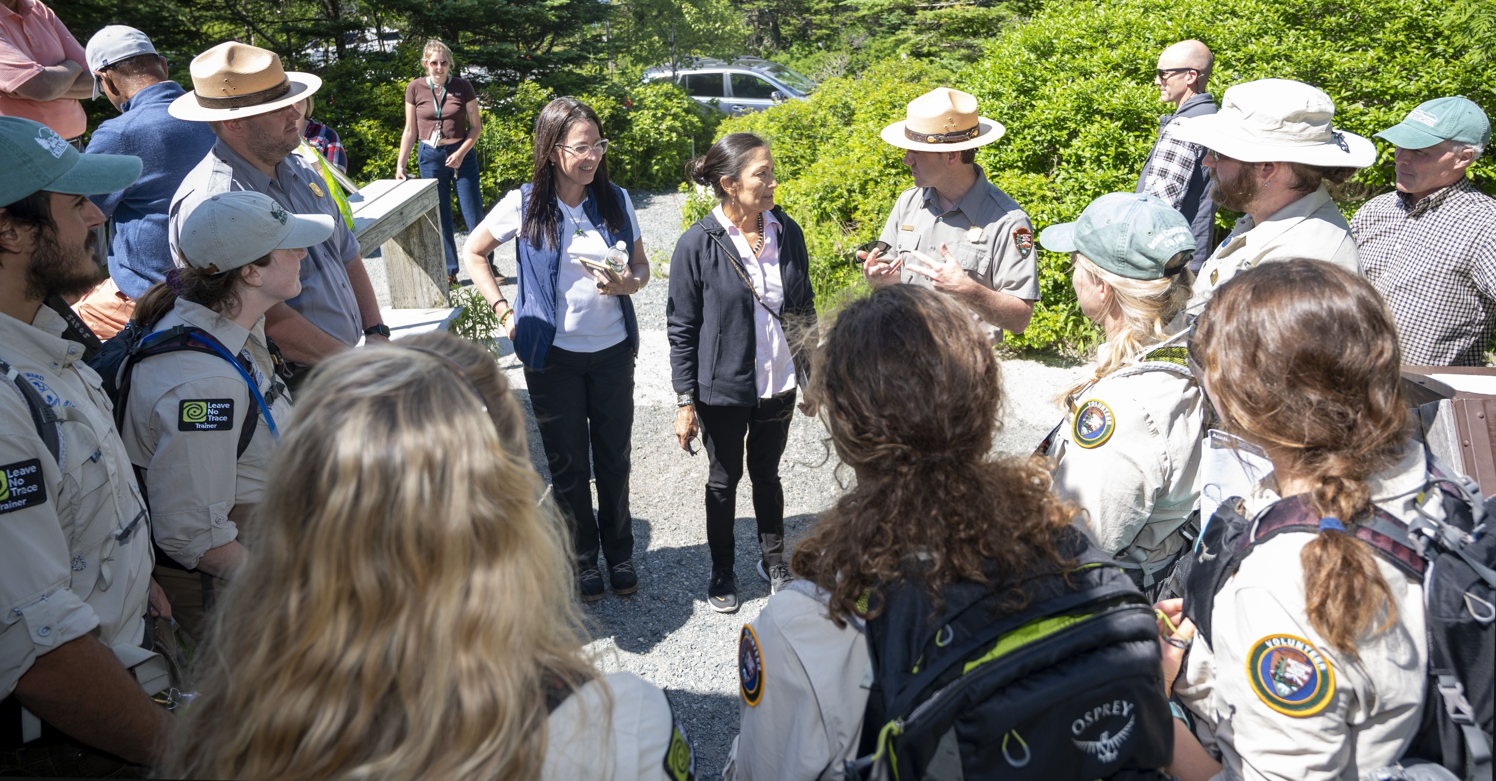 Woman in a black jacket is surrounded by a dozen people in uniform along a gravel pathway fringed with dense shrubbery.