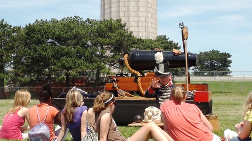 A group of students sit in the grass listening to a man dressed as a War of 1812 infantryman, blue coatee with red and whit trim and stovepipe shacko with a white plume, kneels with various cannon shot in front of him on a table. A carronade, naval cannon is behind him.