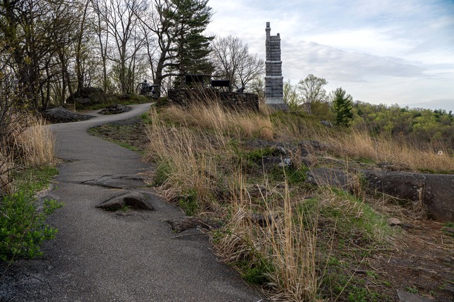 A paved path leads up a to stone monument near a paved platform with a rock wall. To the left of the top of the path are cannon and another stone monument. Large boulders and tall grasses scatter throughout the area.