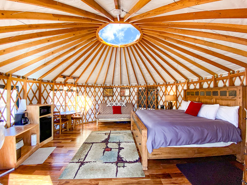 King size bed, mini fridge, microwave, and coffee station. Round yurt structure. It has a clear dome on the ceiling, exposed wood beam ceiling and exposed wood lattice wall supports. A couch and rug can also be seen.