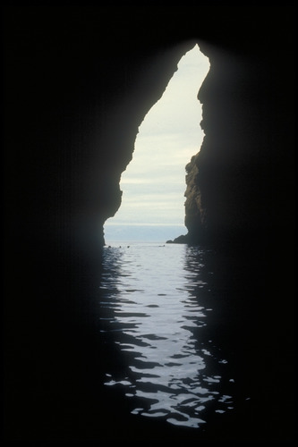 looking through a tall narrow slot opening of a rock wall with water in the foreground and background. 