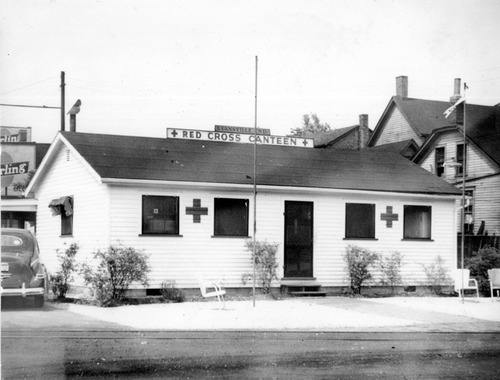 Black and white photo of a white sided one-story building with two crosses on the side and a sign that says "Red Cross Canteen." 