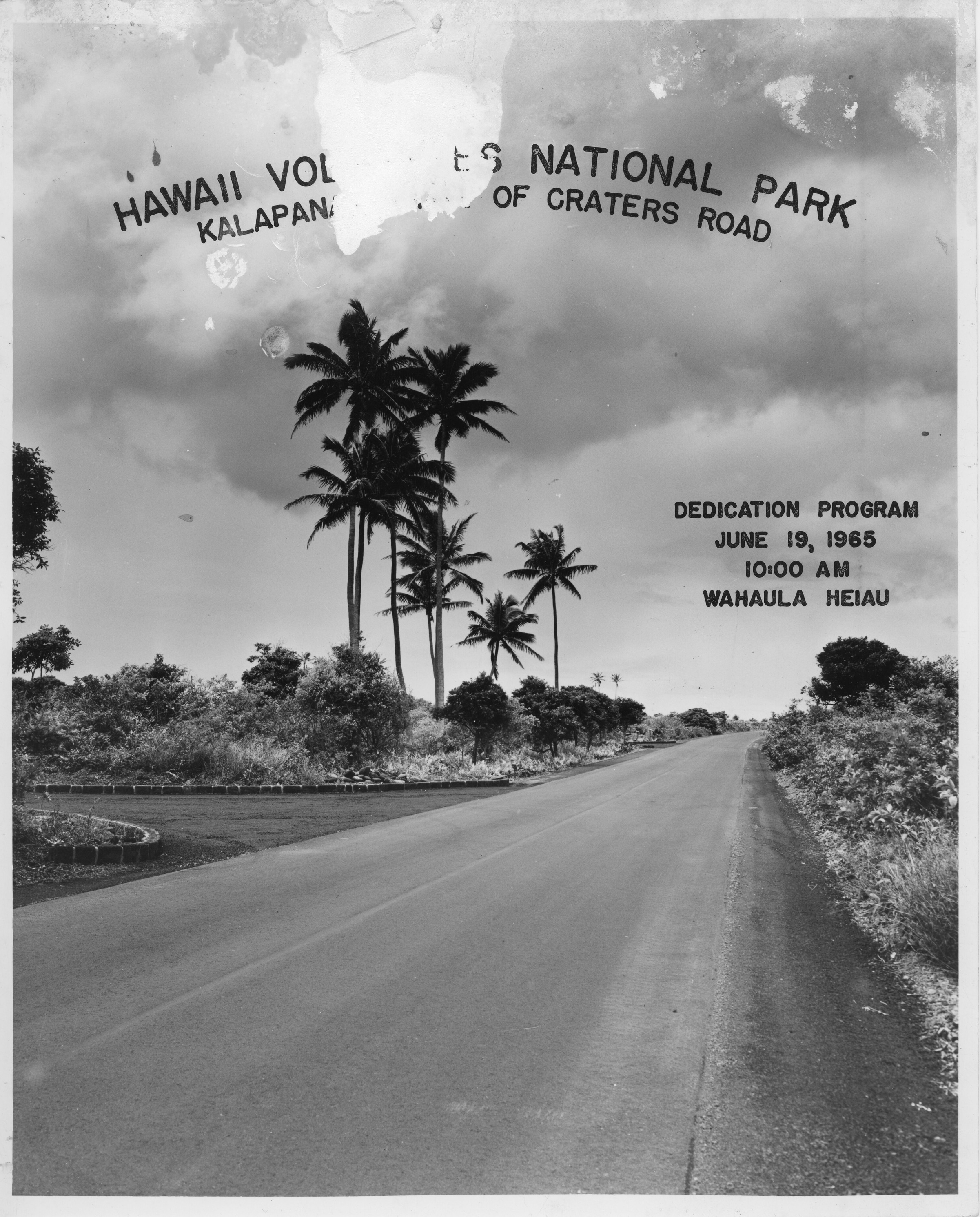 A black and white image of the cover of Hawaiʻi Volcanoes National Park Kalapana Extension of Chain of Craters Road dedication program. The background image of the program cover is a long road that extends from the bottom of the image up to the center of the image. Towards the center of the image there is an off shoot on the road heading towards the left side of the image. Various plants can be seen bordering the road including palm trees, bushes, other trees, and tall grasses. There is a large cloud in the sky located in the top half of the image. On top of the cloud, "HAWAII VOLCANOES NATIONAL PARK KALAPANA EXTENSION OF CRATERS ROAD," is written across it. On the center right side of the image it reads, "Dedication Program June 19, 1965 10:00 AM Wahaʻula Heiau." There is heavy water damage and stains along the top of the image that has cut off most of the word "Volcanoes" in the title.