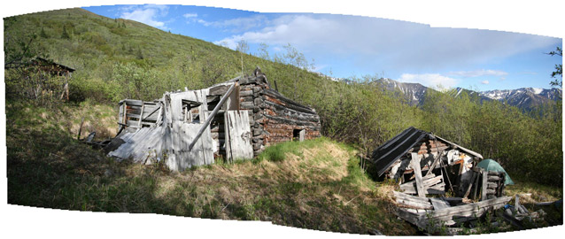 Three log structures, built on terraced terrain with mountains in the background, are falling down. 