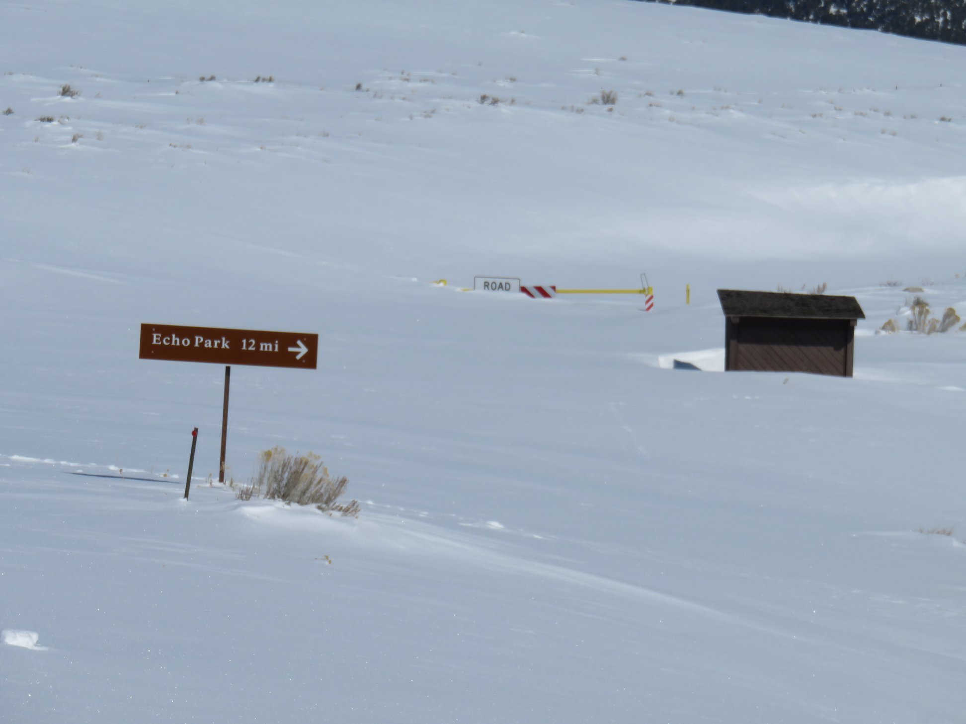 This photo is taken from the location of the Echo Park Road turnoff along Harpers Corner Road, but neither road is visible. Both are buried under several feet of snow. An information kiosk, road sign for Echo Park, and a "road closed" sign peek out of the snow.