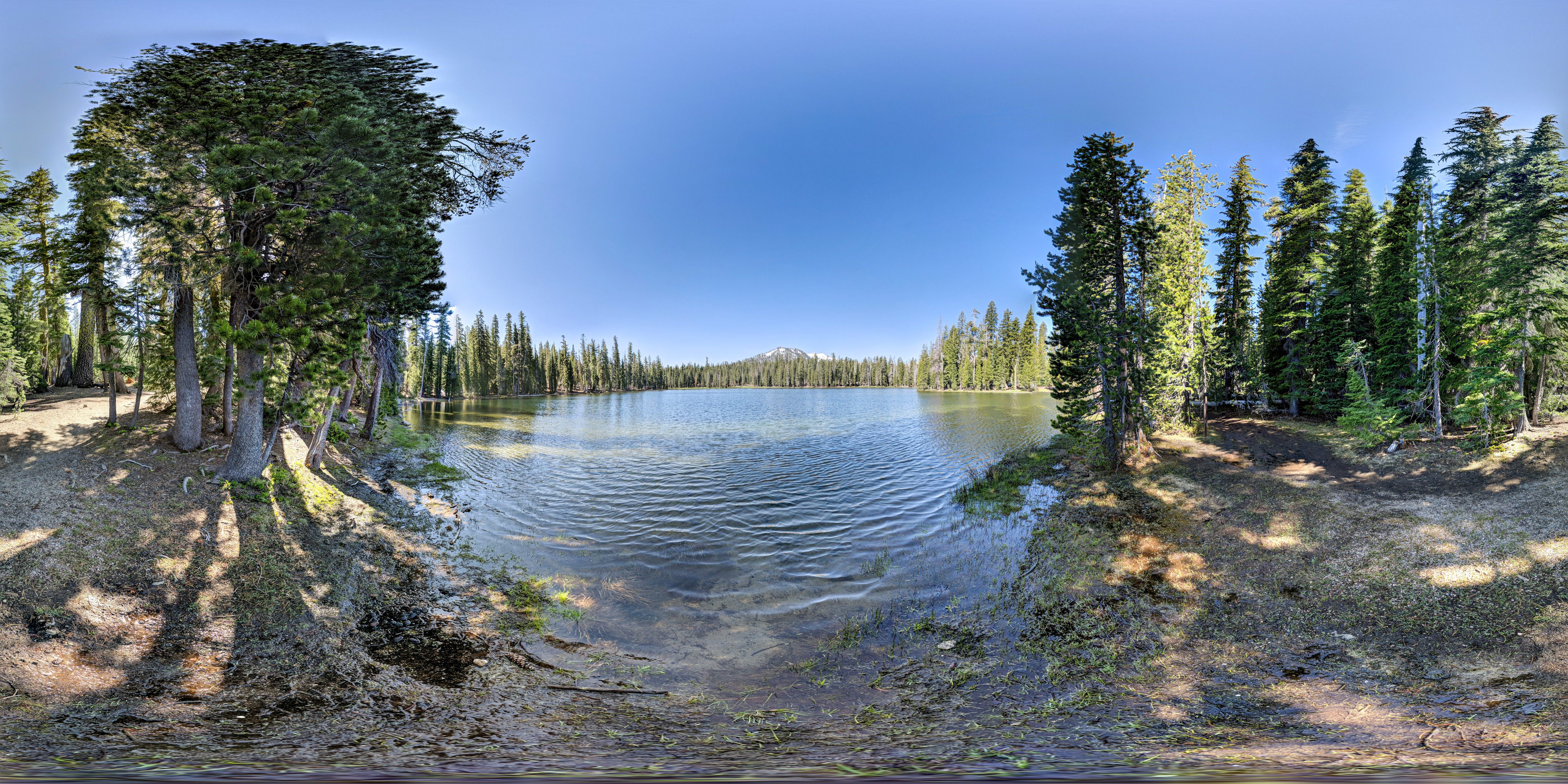 A 360-degree color photo of a lake surrounded by conifer tress. Two snow-capped mountains visible in the distance.