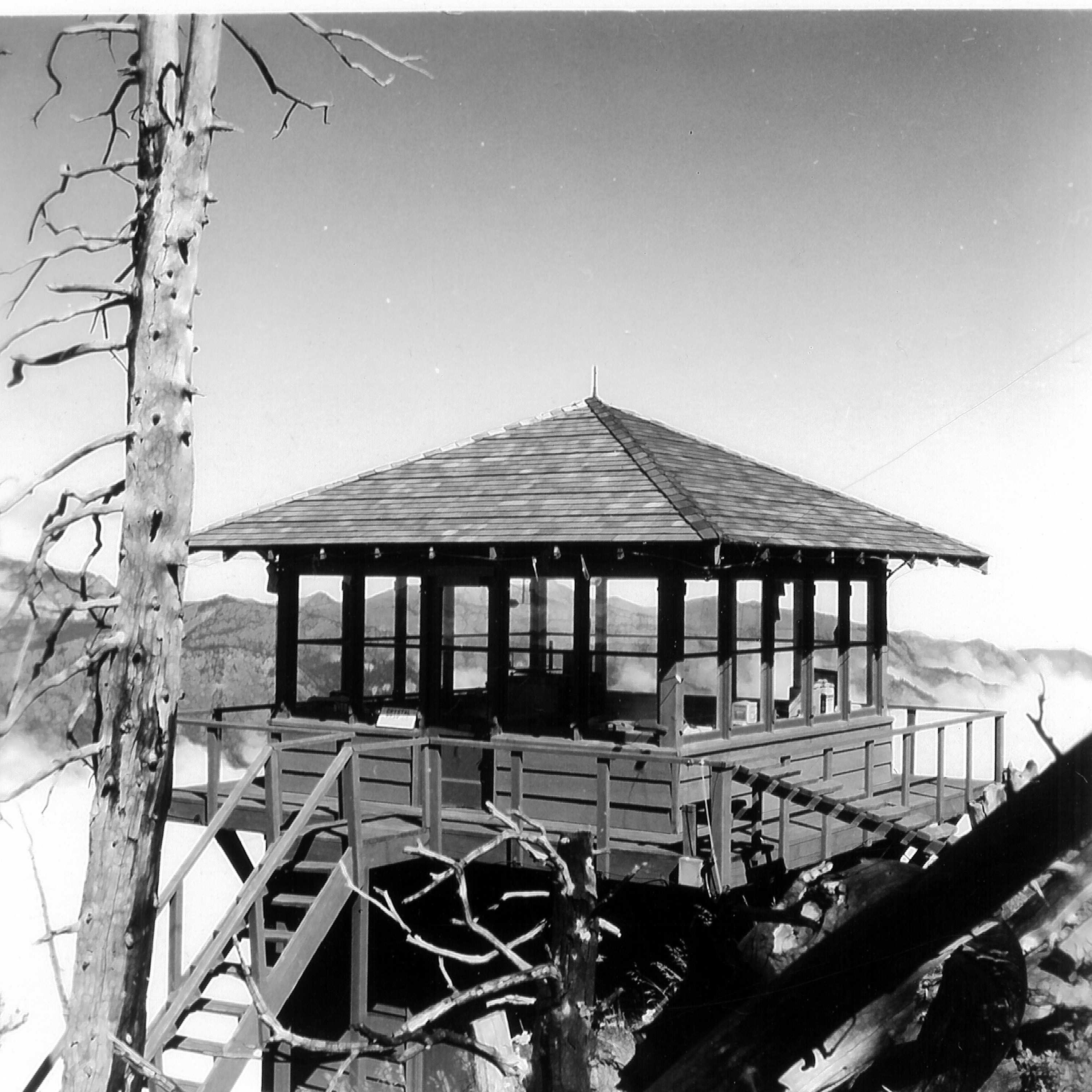 A black and white photo of a square fire outlook building overlooking mountain ridgelines. 
