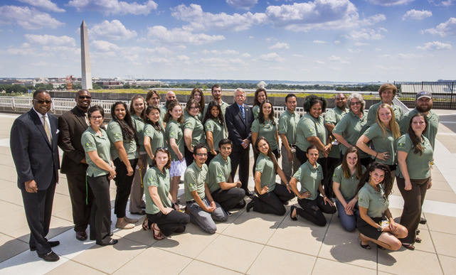 a group of 20 young adults in green polos pose for a photo outside with the Washington Monument in the background. 