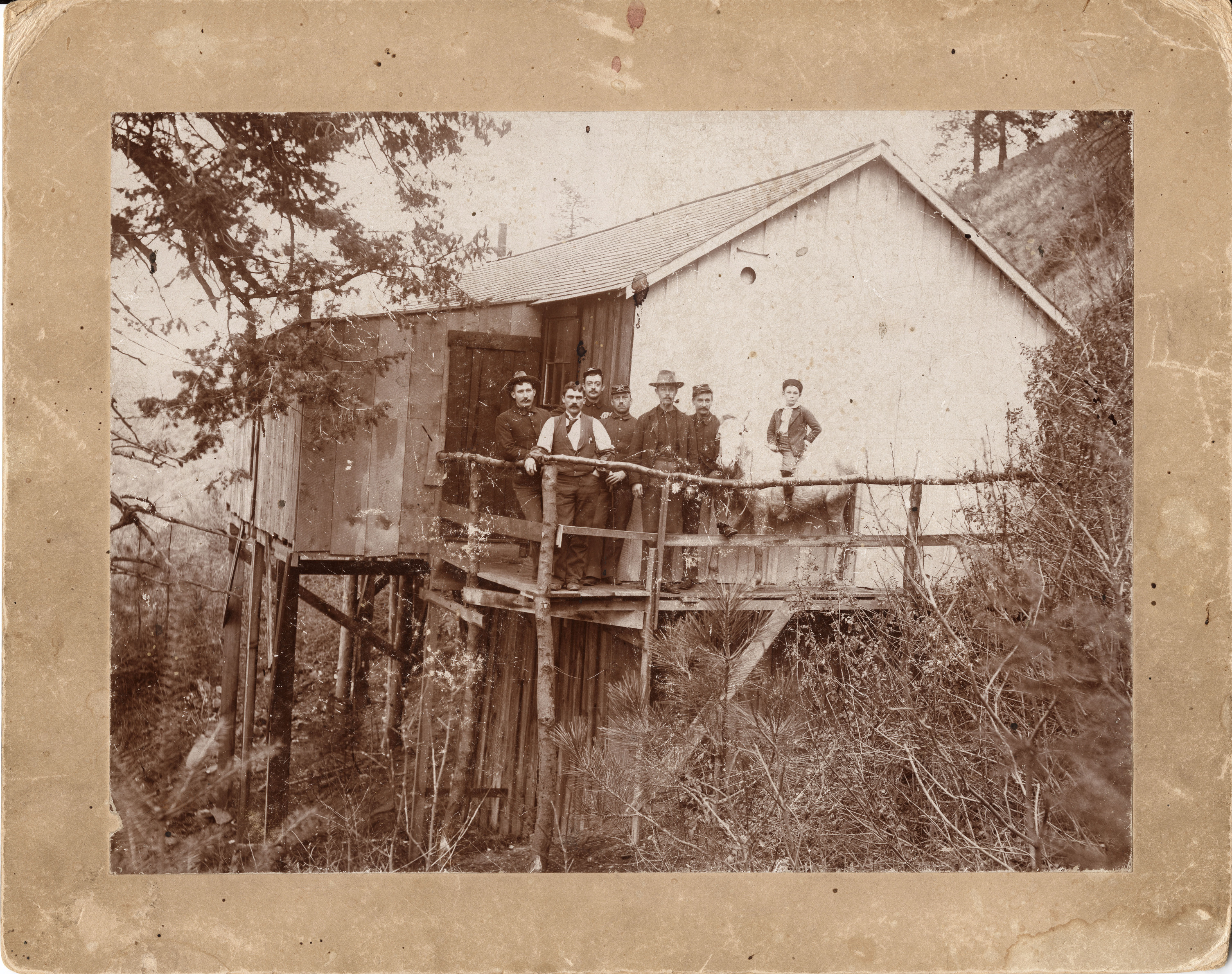 Black and white photograph with tan border of several men and a boy on a horse on the porch of a stilted building on a hillside