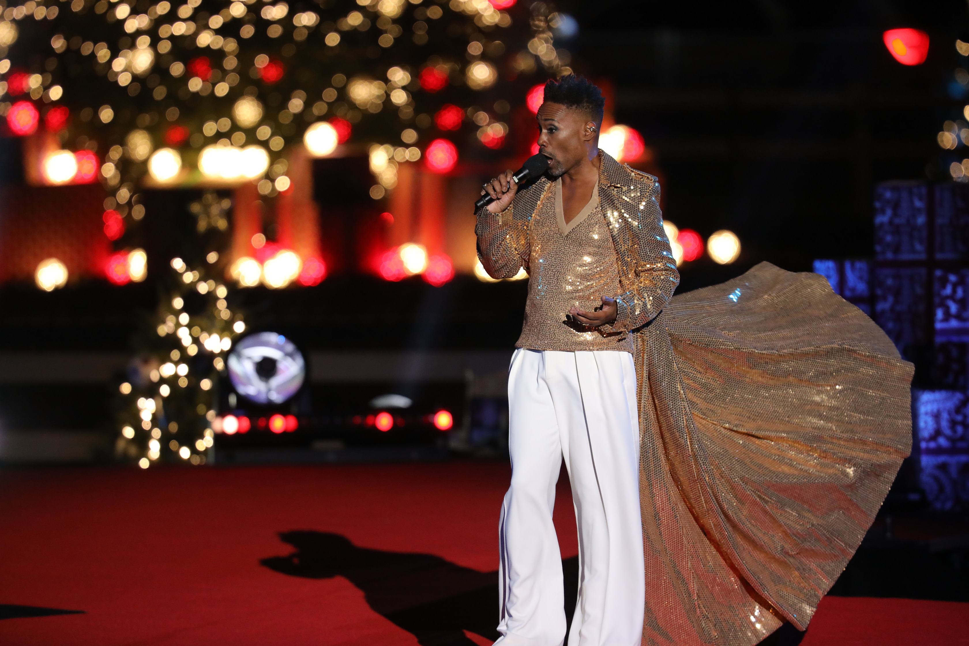 Billy Porter sings onstage. He is wearing white pants and a sparkling gold floor-length jacket that flows behind him.