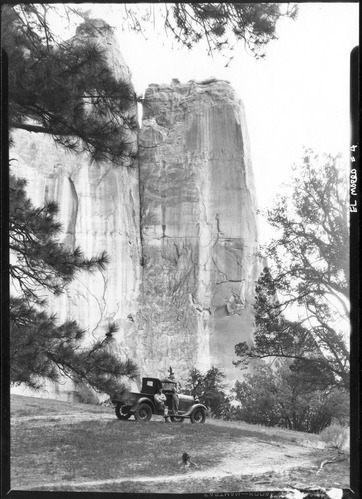 A black and white photo of a man sitting with his legs crossed on a 1920's style vehicle while a young girl stands on the fender, the huge rock face of El Morro Towers behind them.