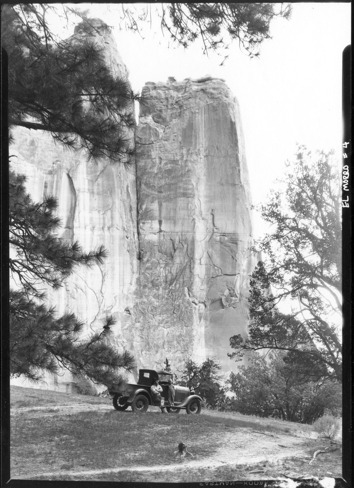 A black and white photo of a man sitting with his legs crossed on a 1920's style vehicle while a young girl stands on the fender, the huge rock face of El Morro Towers behind them.
