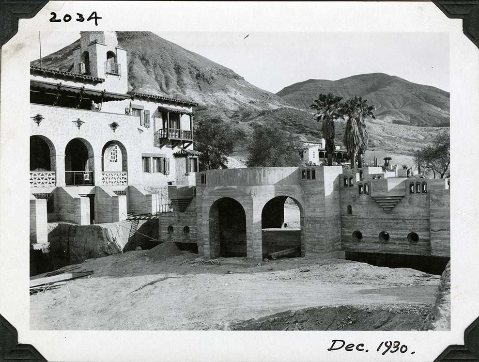 This is an historic black and white photograph from the Scotty's Castle Historic Photograph Collection, Death Valley National Park of dirt road and excavated area foreground. Middle ground is mostly completed arched bridge with round platform over future pool area. Ladder stretches from bridge over trench to area between flying buttresses that will become steps. Spanish style elaborate structure on left and in the distance. Three tall palm trees and desert hillsides in background. Inscription in black ink along upper and lower border.