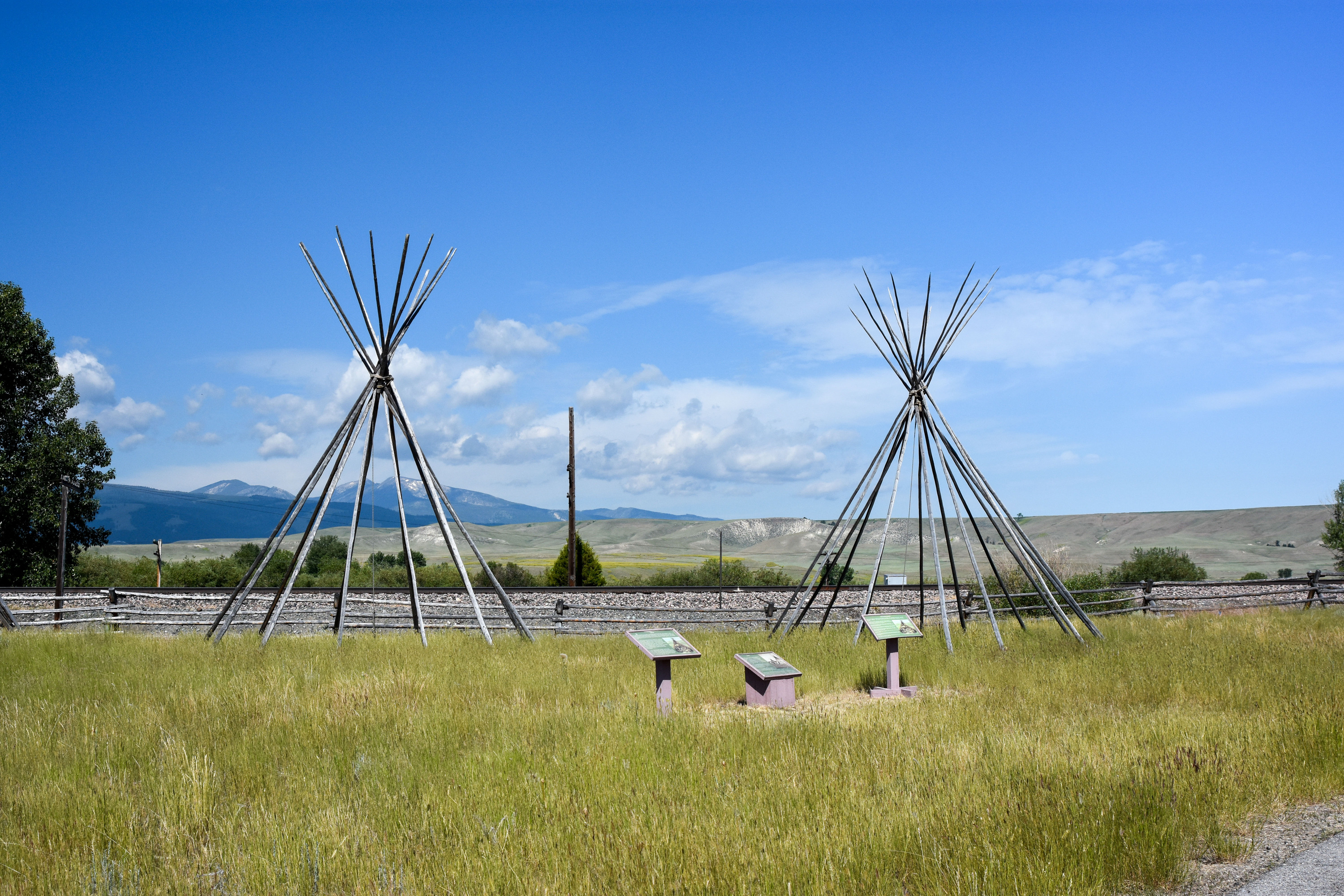 Two tipi structures of poles arranged in a circle and attached at the top in a grassy area beside a set of interpretive markers. 