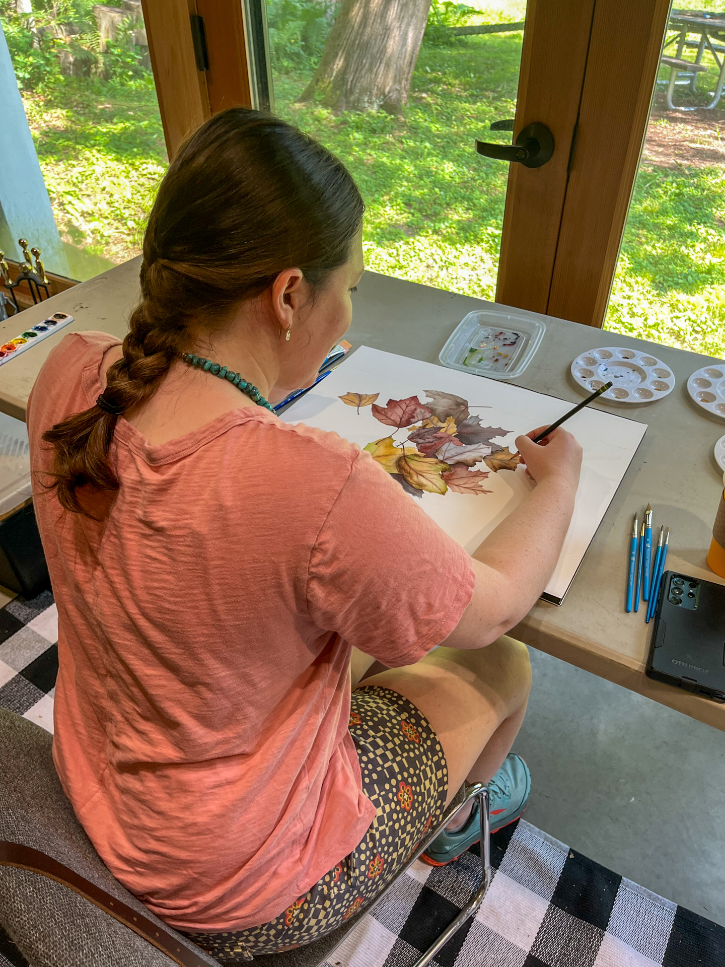 over the shoulder of an artist painting fall leaves in watercolor in front of sunny window