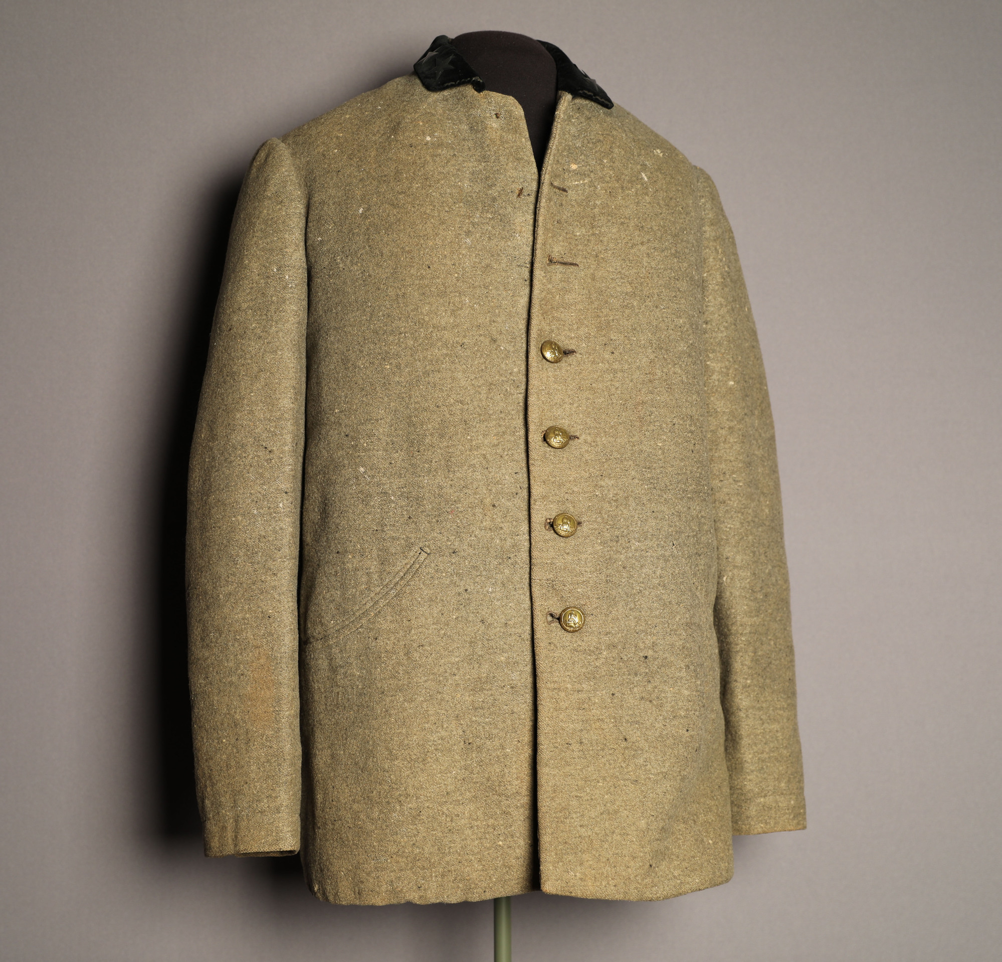 Color photo of the front of the uniform coat of Colonel Simeon B. Gibbons. The coat is khaki colored and made of a woolen material, with a collar and full-length sleeves. The collar is black and made from a velvet-like material. Down the front of the coat are seven buttons, the first three of which are unfastened. The buttons are circular gold metal and have a small design on them.