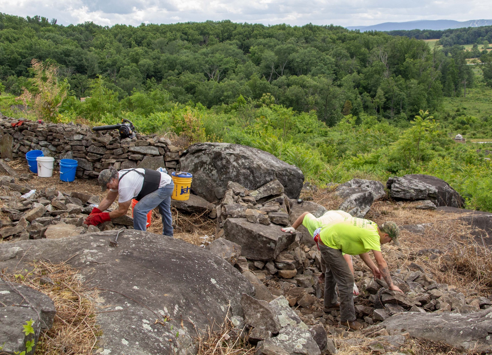 Three stone masons work to pick out appropriate small rocks to complete the restoration of a historic stone wall, with monuments and trees in the background.