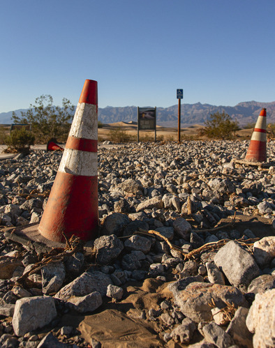 Orange cones sit atop softball sized rocks that cover a parking lot leading to a information panel with sand dunes in the background.