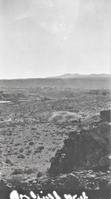This is an historic black and white photograph from the Scotty's Castle Historic Photograph Collection, Death Valley National Park of and expanse of desert landscape with some mining activity in middle ground and town site in the distance. Taken from hillside of very rough rock and rubble. Circular tailings with square shaft in middle ground. More extensive tailings and several structures of probably a milling site. Faded townsite in distance below low mountains. Negative had illegible writing across bottom edge that appears white on print.
