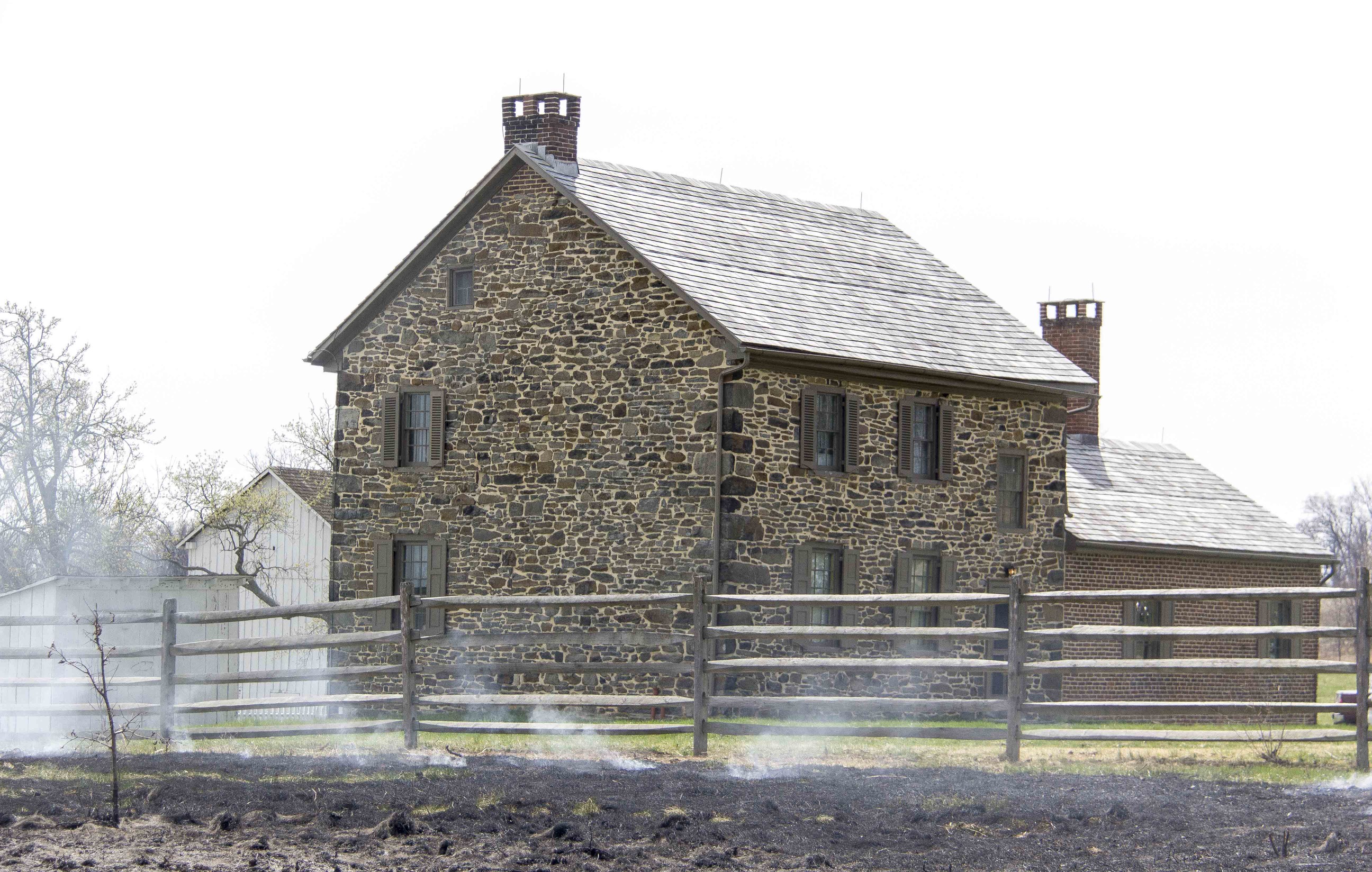 The prescribed fire has moved around and past the Bushman house. The stone house is center in the picture. Smoke is making its way around and past the house. There is a wooden fence. 