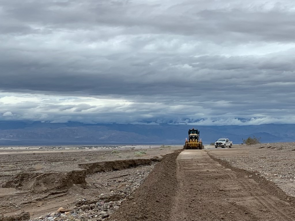 Heavy equipment clears debris from one lane of a paved road in a desert landscape with a park ranger truck and mountains in the background and dark clouds overhead.