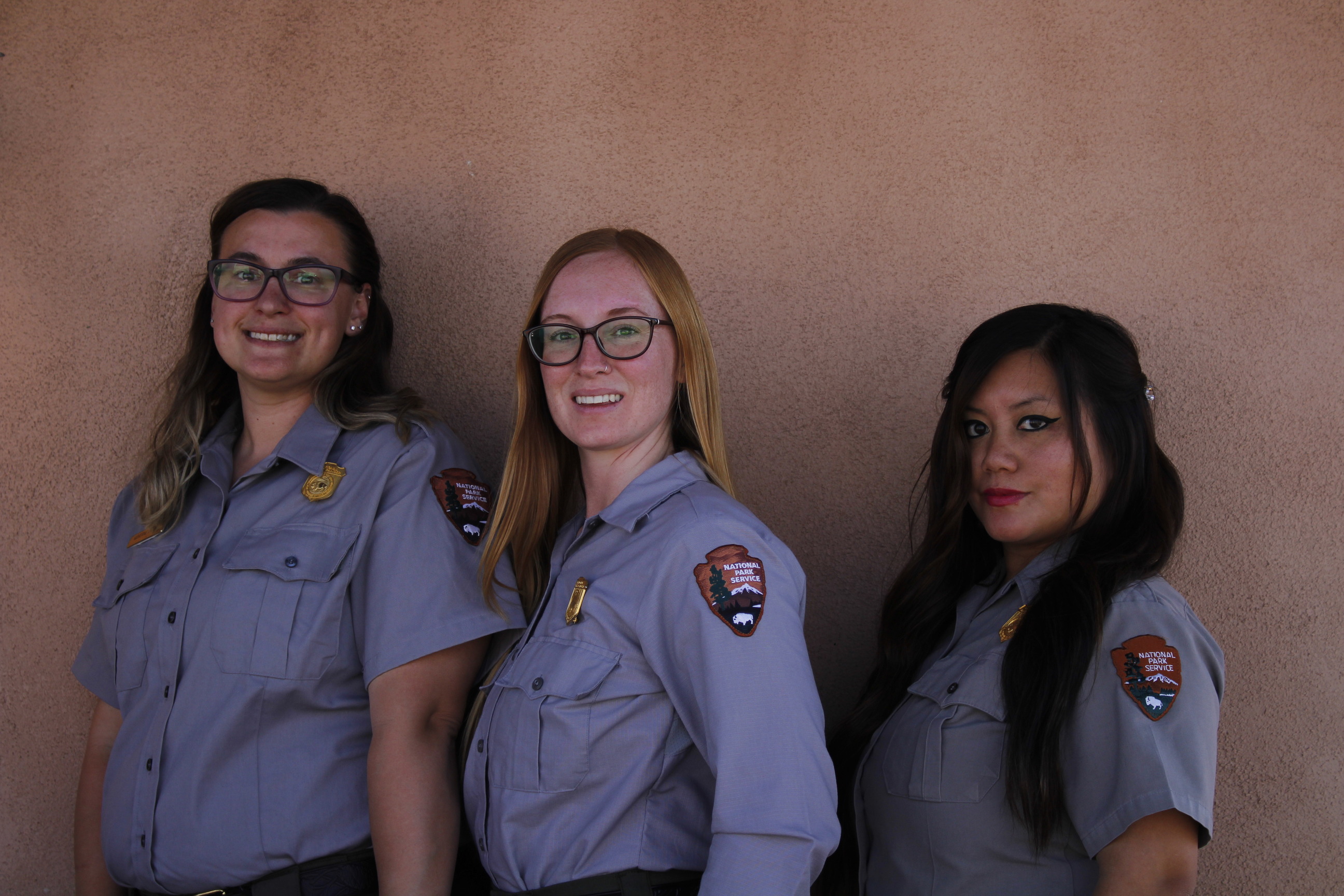 Three women wearing the National Park Service uniform pose for a photo against a tan stucco wall. 