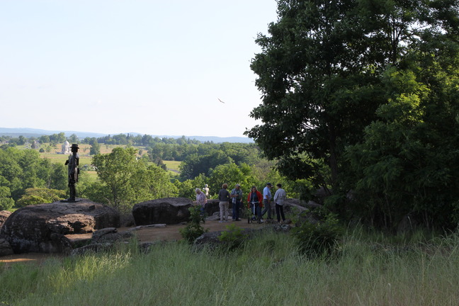A bronze statue stands on a boulder looking out over a hill. People stand in a viewing area to the right. 