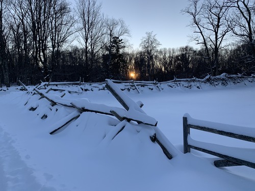 Fresh snow covers the ground and the stones and wooden rails of a fence. Beyond behind bare trees the sun is setting.
