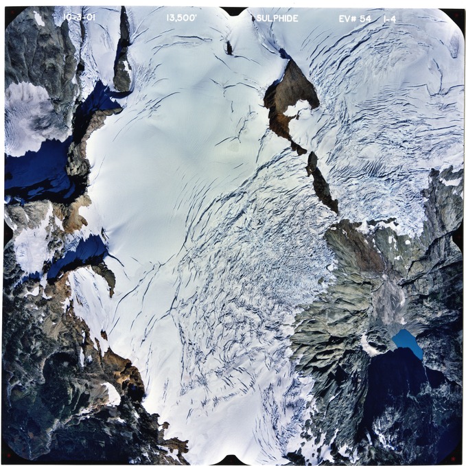 Color aerial photograph of glacier and alpine lake on mountain peak.