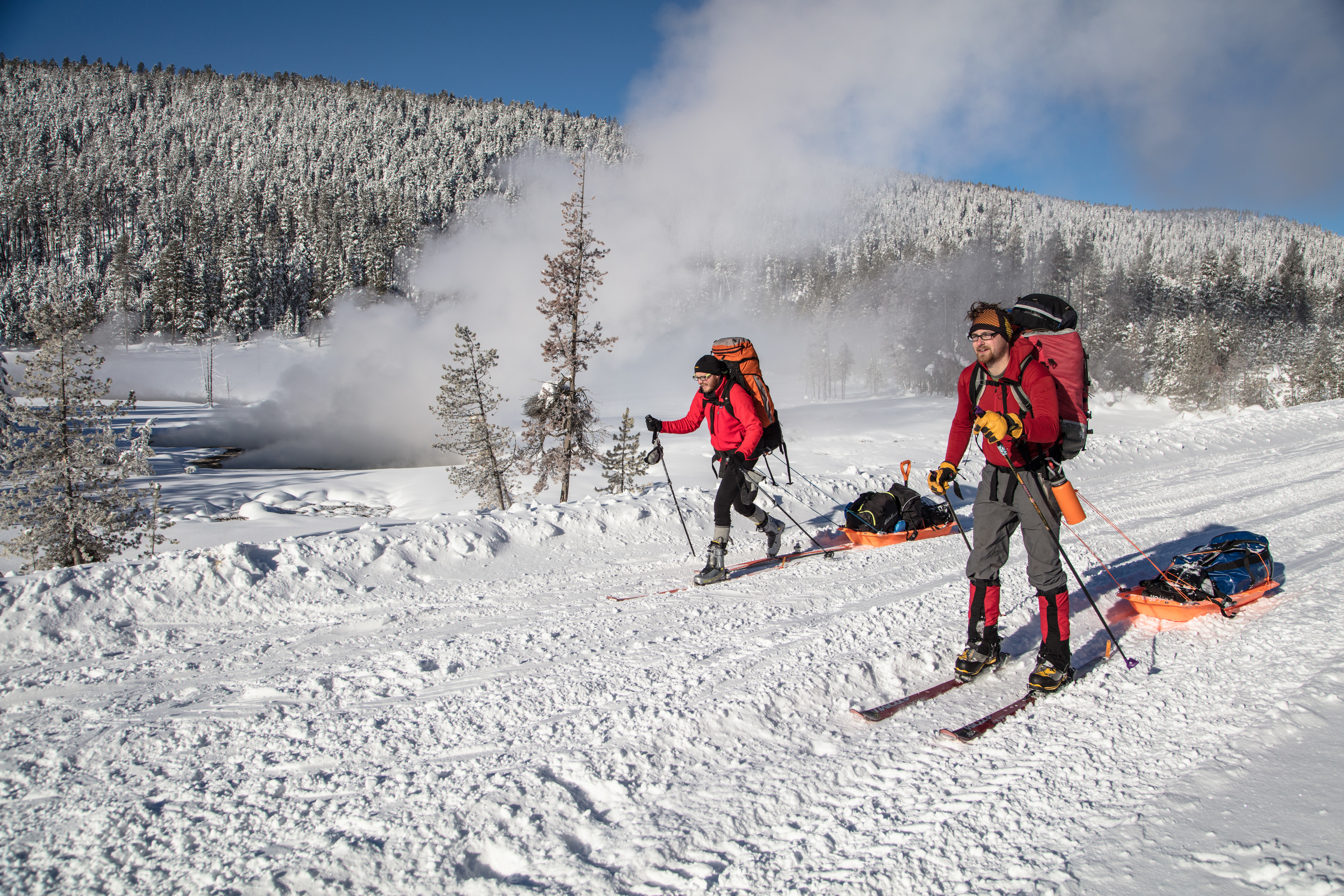 Two skiers with backpacks pulling sleds on a groomed road with steam rising in background