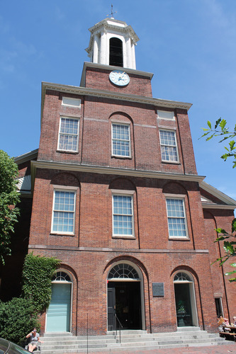This photograph shows the Charles Street Meeting House, originally a white Baptist Church. The church building sits in the middle of the photograph. It is a red brick building approximately four stories tall and is topped with a white, wooden steeple which is at the front of the roof and ends in a curved triangle at the top. The steeple has at least two large arch openings in it. On the first floor there is a wooden door for the entrance into the structure. There are three windows facing the street and one window on the side of the building on the second and third floors. These windows have many panes of glass in them. On the fourth floor there is a clock inset into the area between the floor and the base of the steeple. The Meeting House is surrounded by other red brick buildings on either side. The photograph was taken in the spring and the trees that are in the foreground on the left and right side of the photograph show early white and light green buds. The sky is light blue and has no clouds.