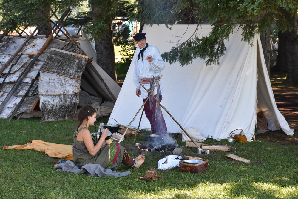 Reenactors cooking near fire, canvas and birch bark shelters.