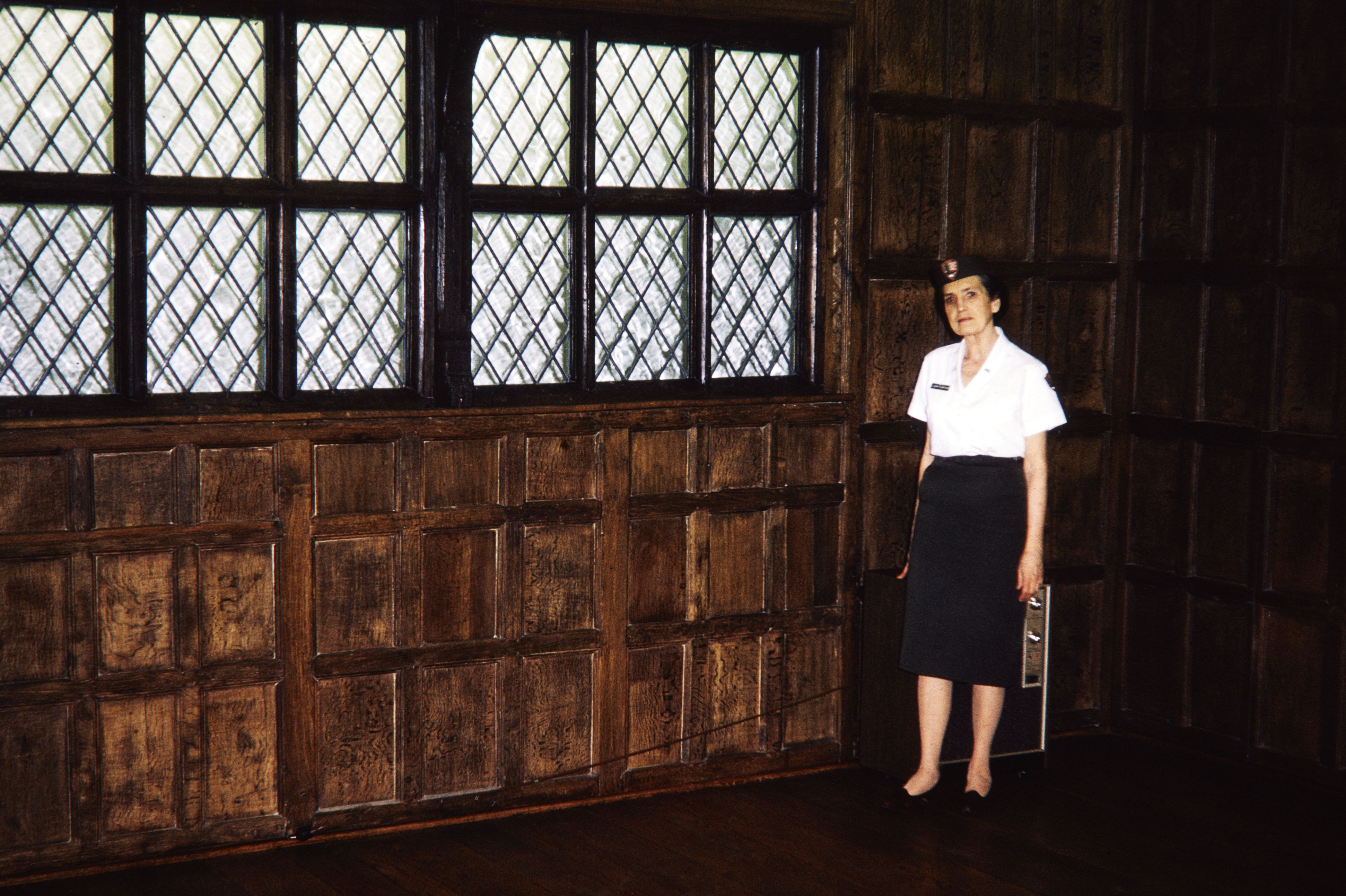 Woman in NPS uniform stands in a room with dark wood paneling and windows. 
