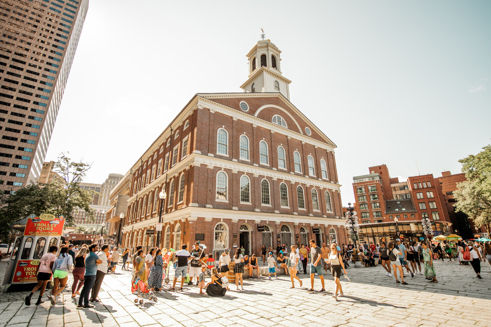 Busy square area in front of a four-story federal-style brick building known as Faneuil Hall. The building has rows of arched windows on each floor and is topped by a pediment with two circular windows on either side of a half-circle window. The building is topped by a cupola with a grasshopper weathervane.