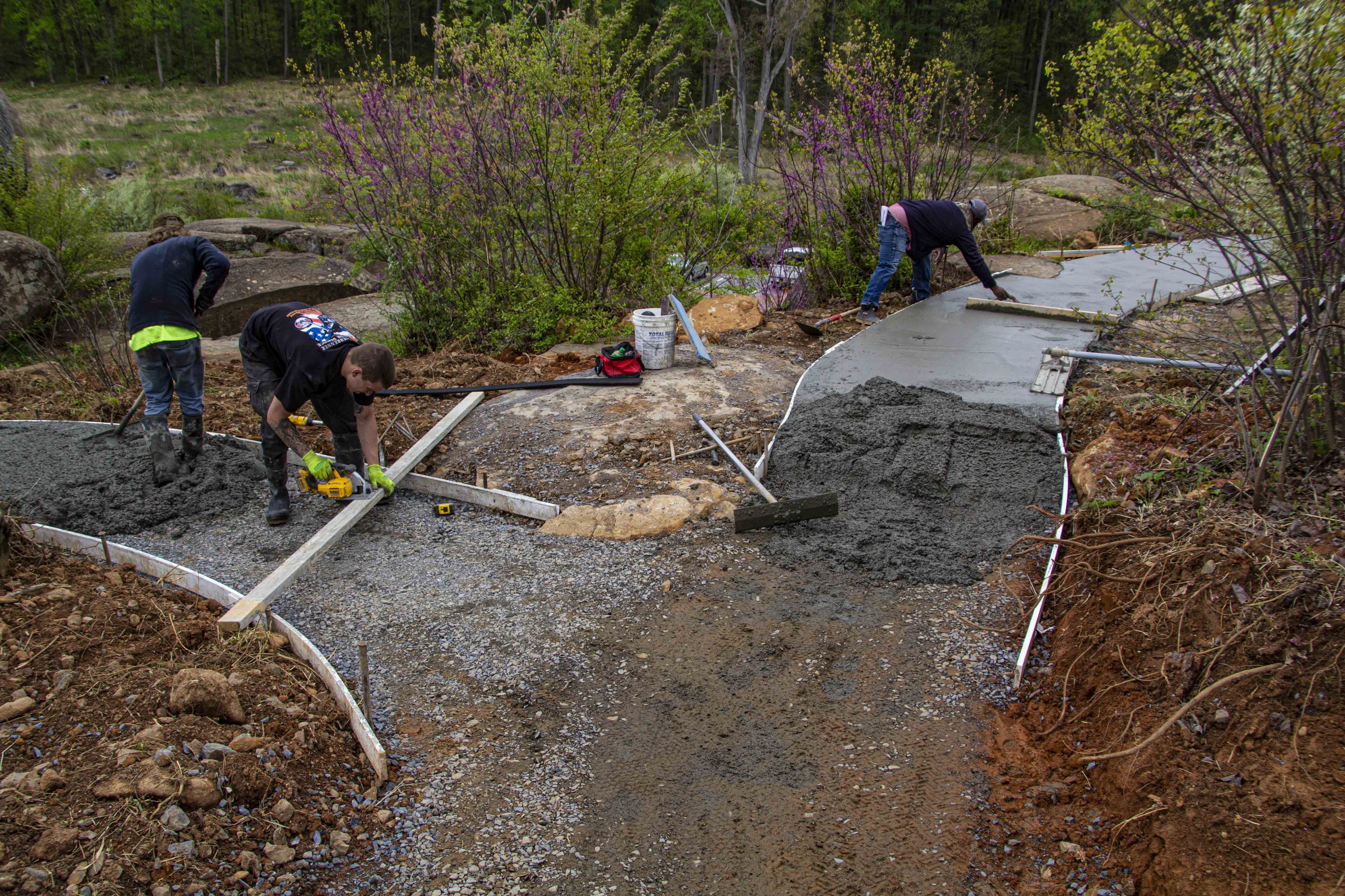 Center-frame is a fork in a partially paved footpath. In the upper-right path of the fork is freshly paved cement with a worker kneeling over to examine it. In the path to the right is a pair of workers measuring the dimensions of the path and smoothing out the freshly-laid cement. In the bottom-center of the frame is a section of the path that hasn’t had any cement laid yet. There are some construction implements in the crook on the side of the road. 