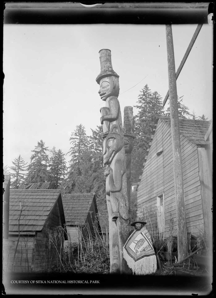 Alaska Native man in Killer Whale Chilkat robe and spruce root hat next to Wolf Pole in Old Kasaan.