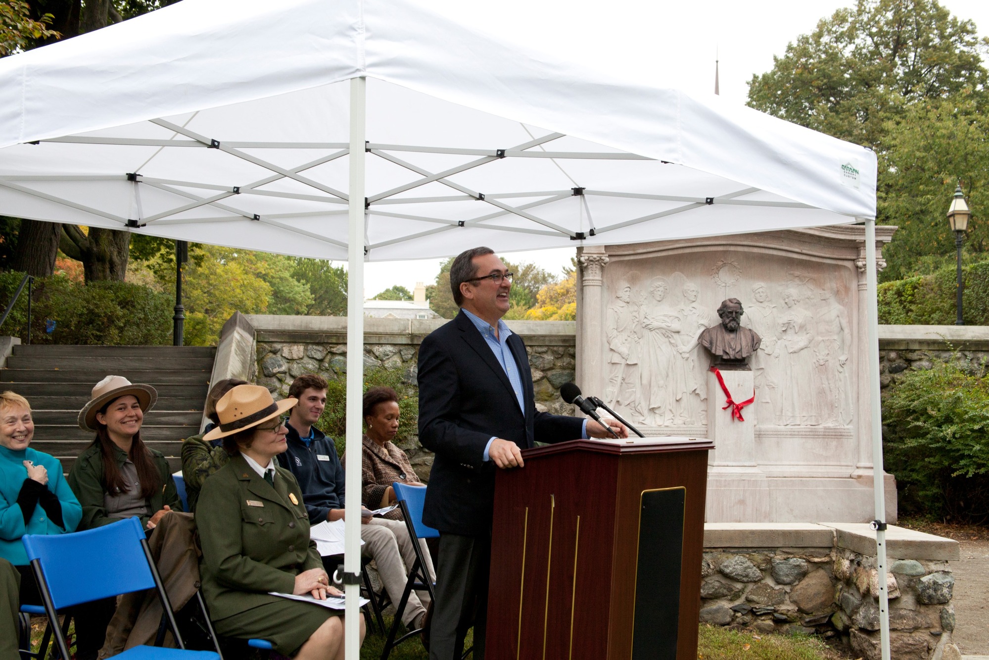 Woman speaks at podium in front of small group seated to left of monument draped with red bow.