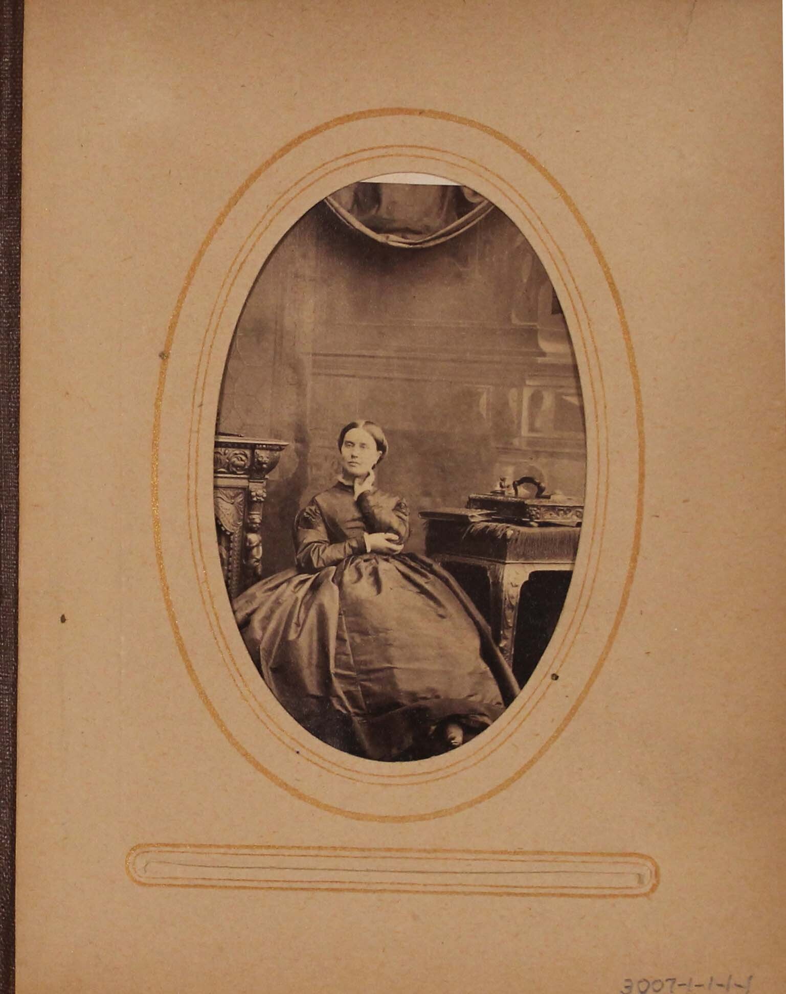 Black and white photograph of woman seated in between fire place and desk.