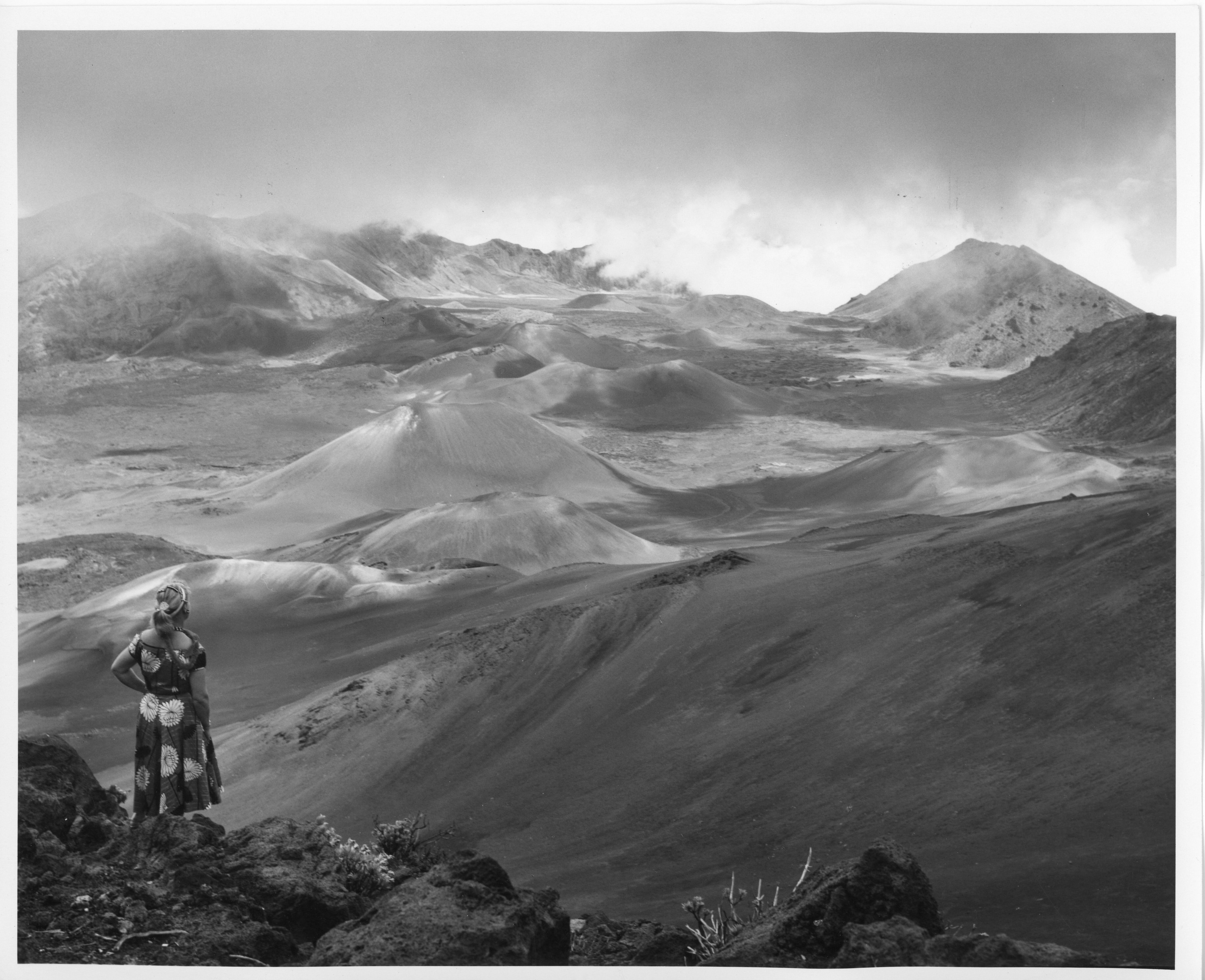 A black and white image of a woman gazing at the cinder cones in the crater of Haleakalā crater. The image shows the woman stand at the left side of the image with her back towards the camera. She is pictured wearing a floral dress and her hair is tied back with a head band. She is also wearing a fabric lei that is hanging on the right side of her shoulder. She is standing gazing at the view with her left hand on her hip. She appears to be standing on rocks and near the edge of the crater. There are large rocks on the ground she is standing on with some bare plants. The view in front of her is the cinder cones in the distance. The cinder cones appear to look smooth and pointy at the top. Below the cinder cones there is hardened lava and rocks. There are mountains to the left and right of the image in the background. The clouds in the sky appear to be sitting over the mountains in the distance.